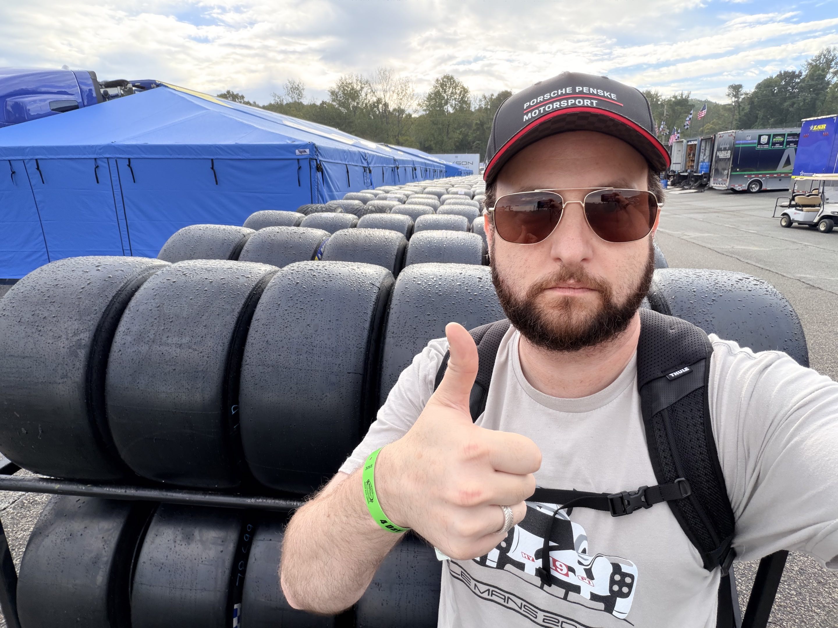 A man in a race car t-shirt, race car hat, and sunglasses wearing a wristband and a backpack giving a thumbs up behind an endless row of racks of slick racing tires